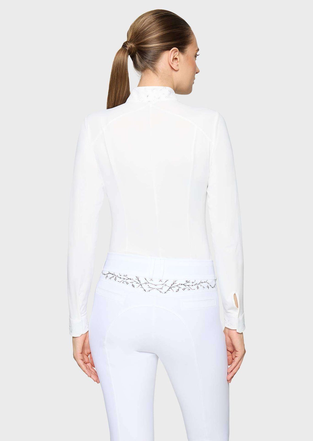 A woman shown from the back wearing a white long-sleeve mock-neck equestrian-style top and matching white fitted pants with a decorative silver embroidered waistband.