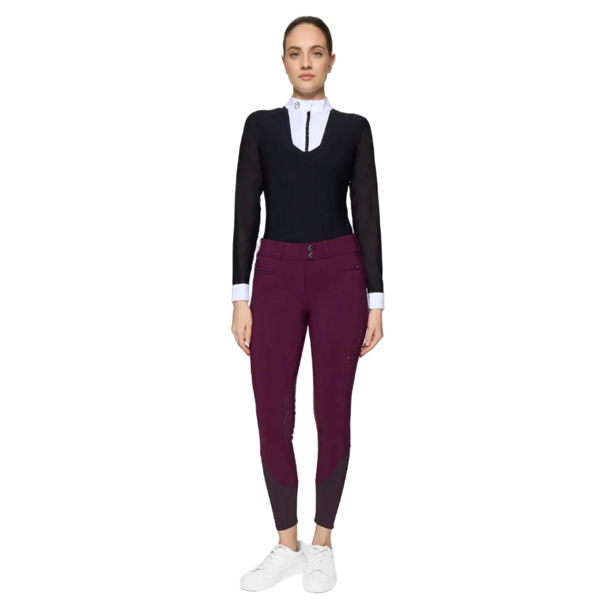 A woman modeling a black long-sleeve equestrian-style top with a white mock neck collar and dark purple fitted riding pants with dark knee patches, paired with white sneakers, against a light gray studio background.