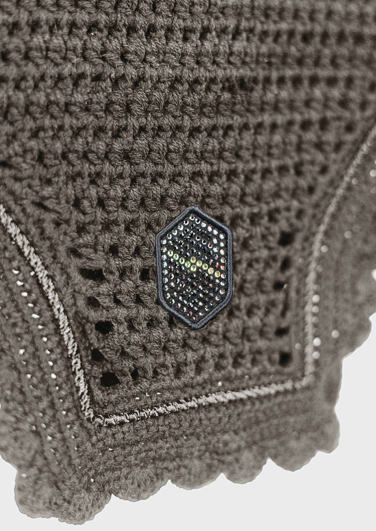 Close-up of a dark brown crocheted textile with a scalloped edge, featuring a small hexagonal metallic logo charm on the front.