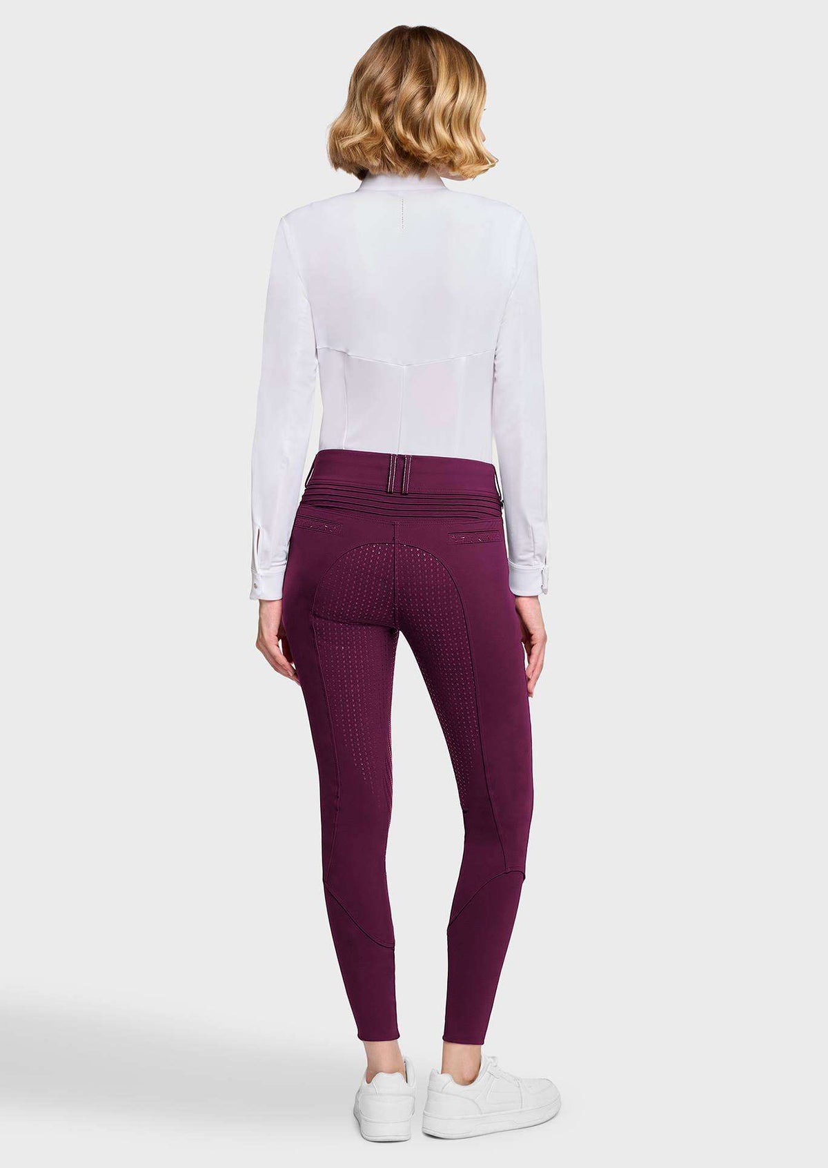 A woman shown from the back wearing dark plum riding breeches, a white long-sleeve shirt, and white sneakers against a light gray studio background.