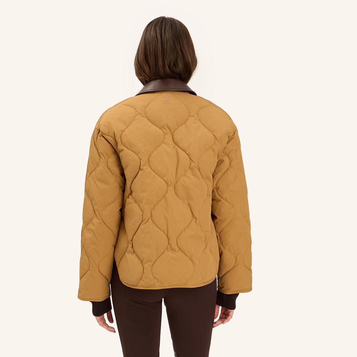 A model viewed from the back wearing a mustard yellow quilted jacket with dark brown trim, paired with dark brown pants and black knee-high boots, against a solid off-white background.