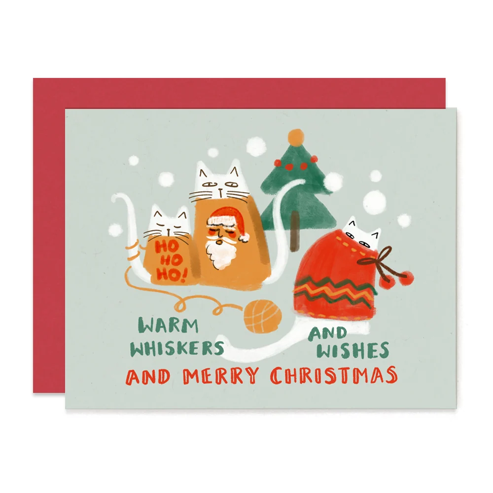 A Christmas greeting card with a red envelope behind it. The card features a light gray background with a whimsical illustration of three cats knitting holiday sweaters and hats, with the text 'WARM WHISKERS AND MERRY CHRISTMAS'.