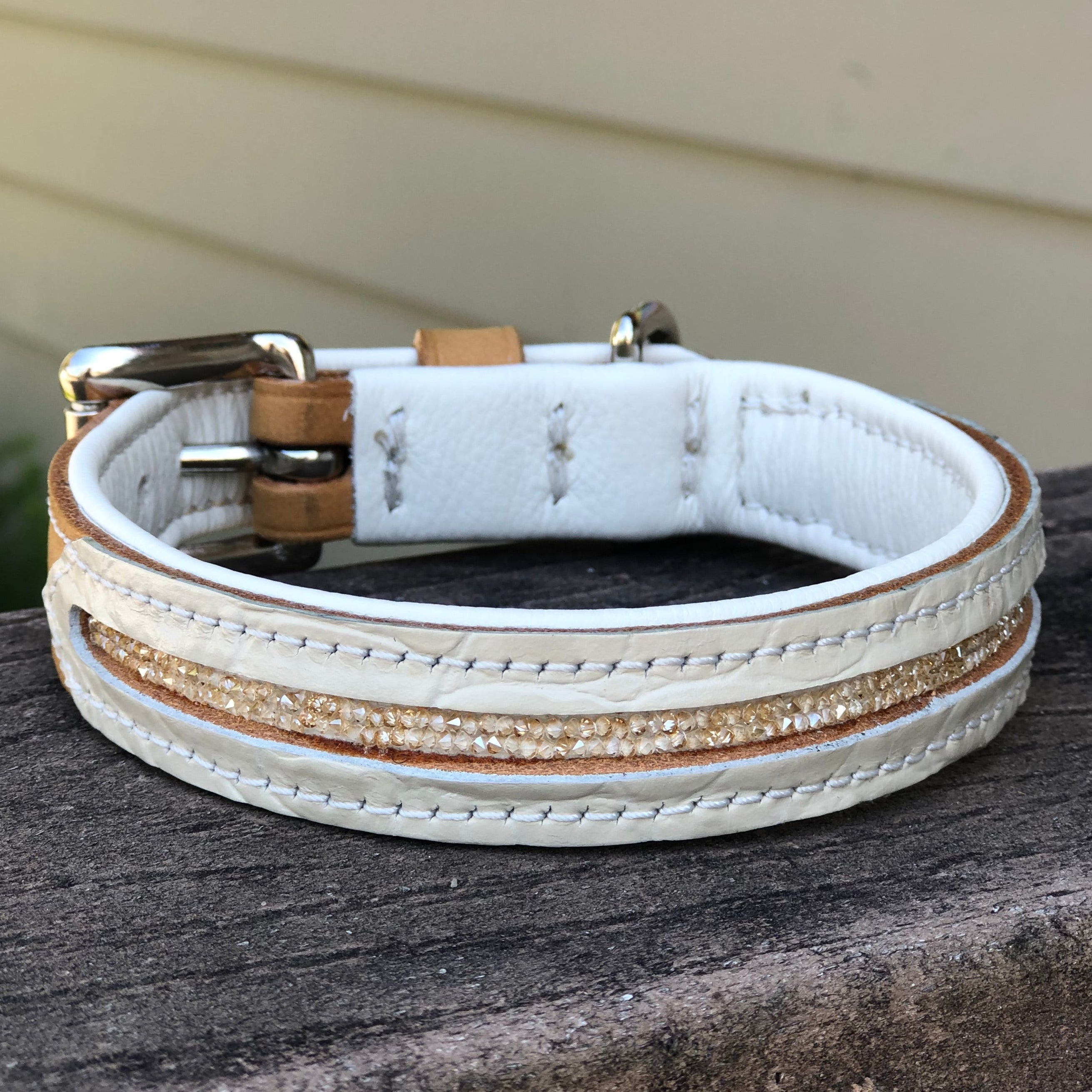 A white leather dog collar with a textured gold glitter stripe down the center and a silver-tone buckle, resting on a weathered wooden surface.