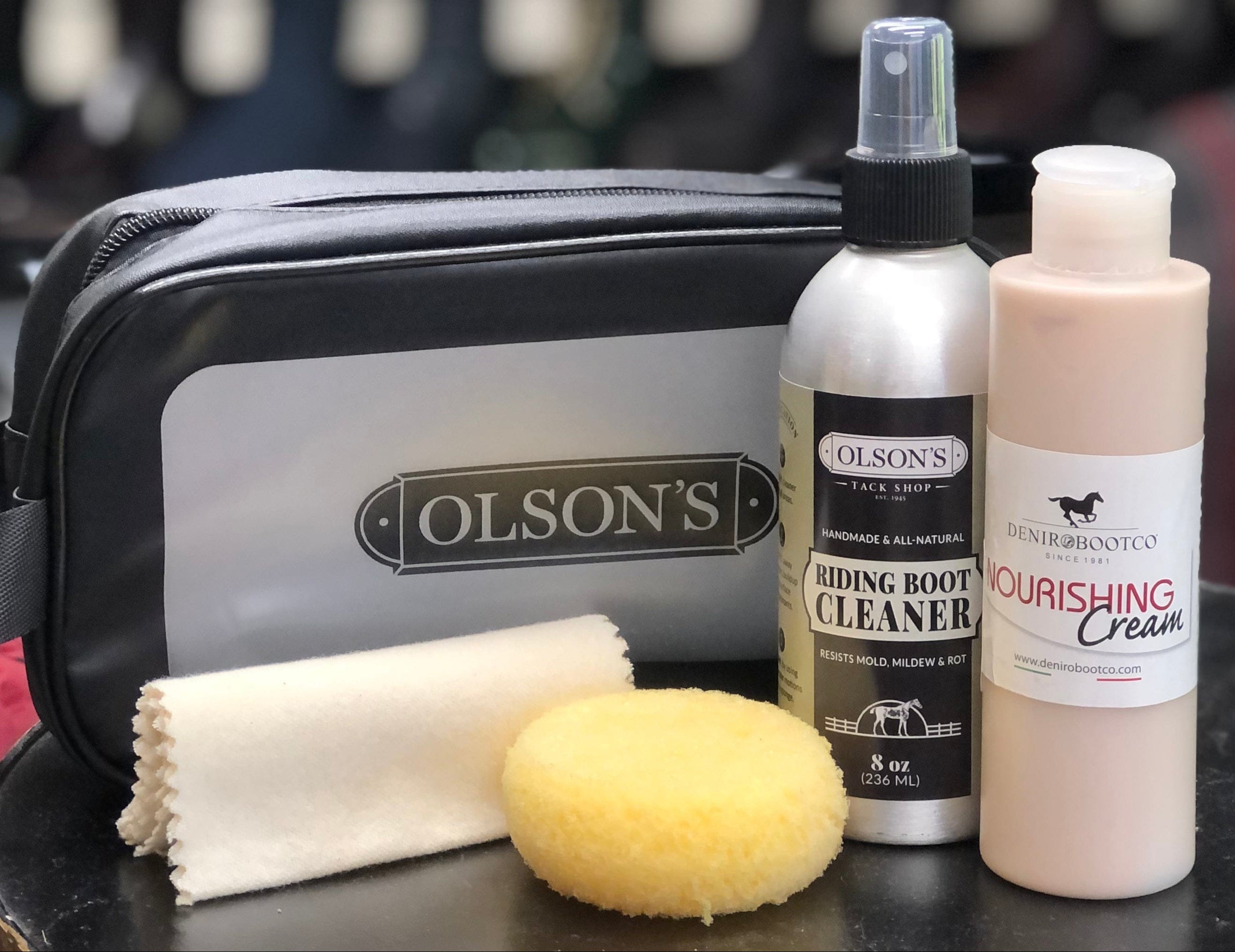 Olson's leather care kit including a spray boot cleaner bottle, cream, and sponge on a table with shoes in the background.