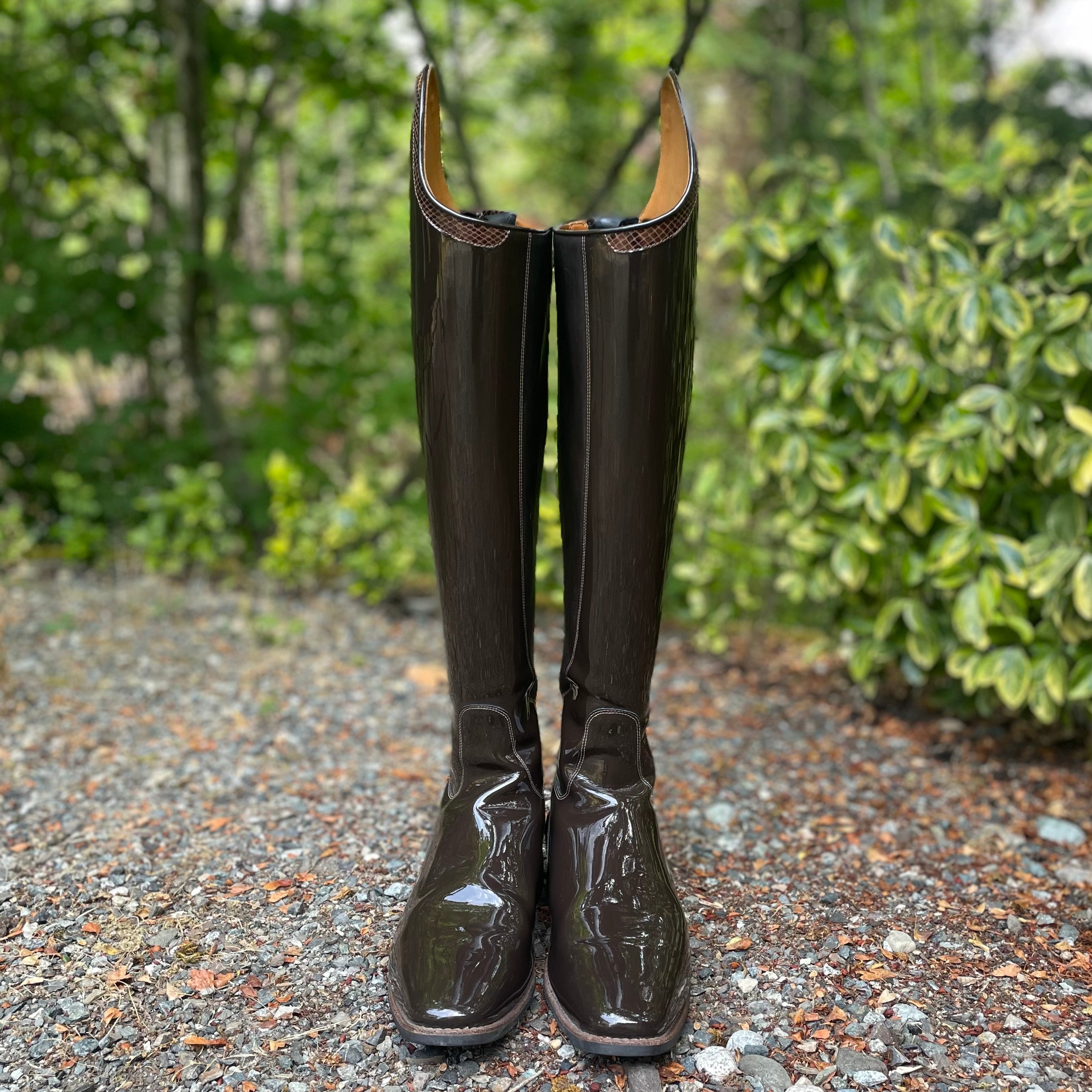 A pair of tall glossy dark brown rain boots standing upright on a gravel path with green foliage in the background.
