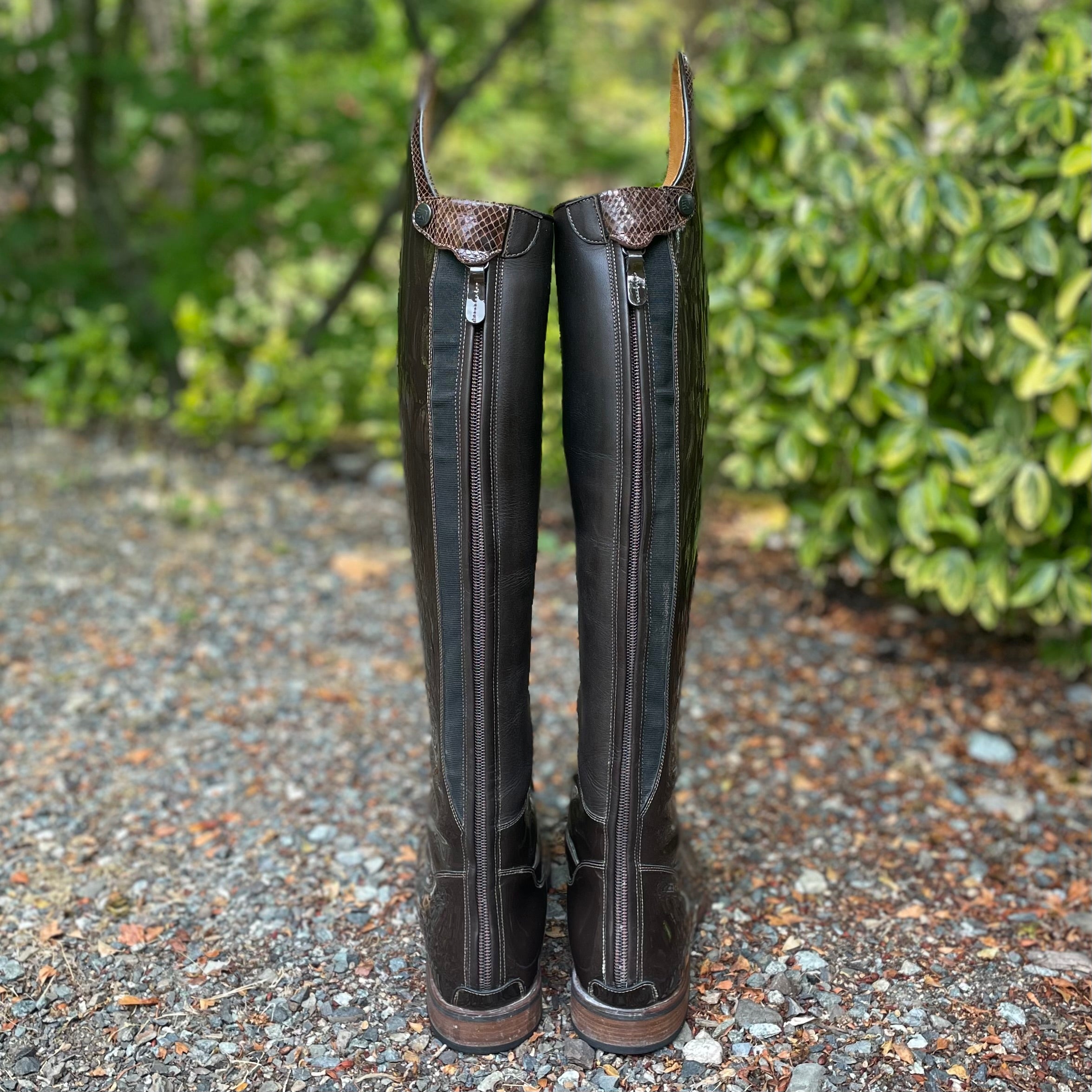 A pair of tall dark brown leather riding boots shown from the back, standing on a gravel path with green foliage in the background.