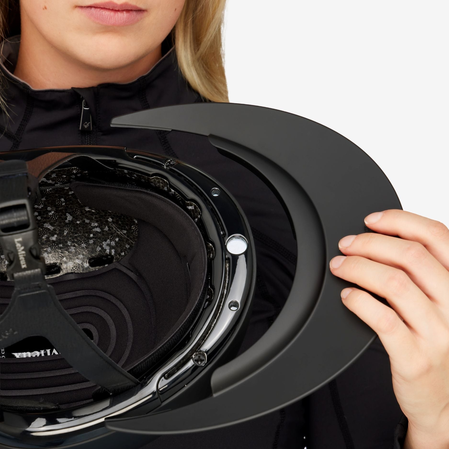 A close-up of a woman holding a black horse back riding helmet with its visor open, showing the interior padding and straps.