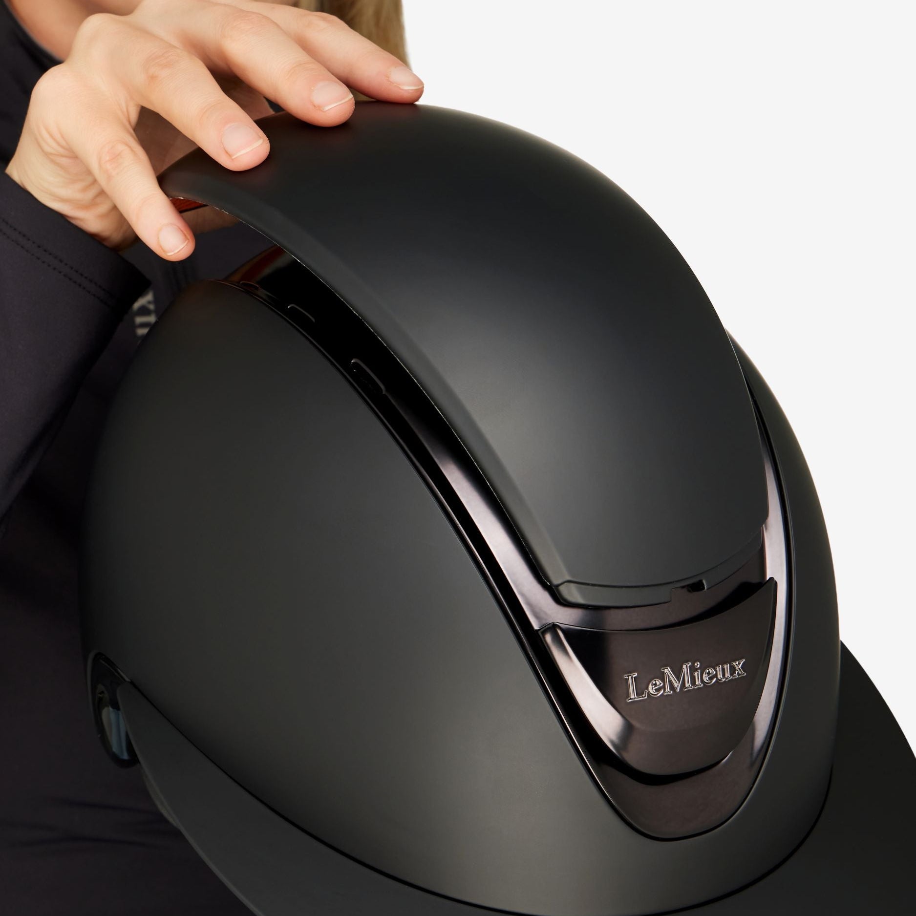 A close-up of a person holding a matte black LeMieux equestrian helmet against a light gray background.
