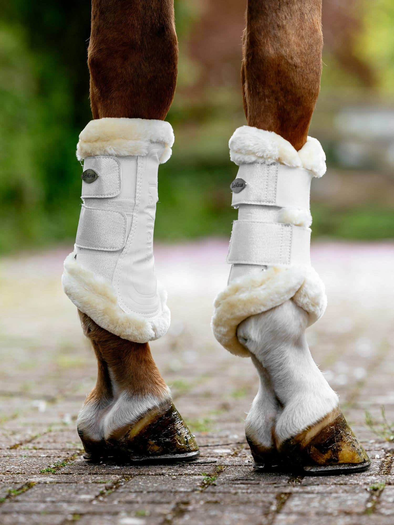 Horse legs wearing white protective boots with fur trim on a brick surface.
