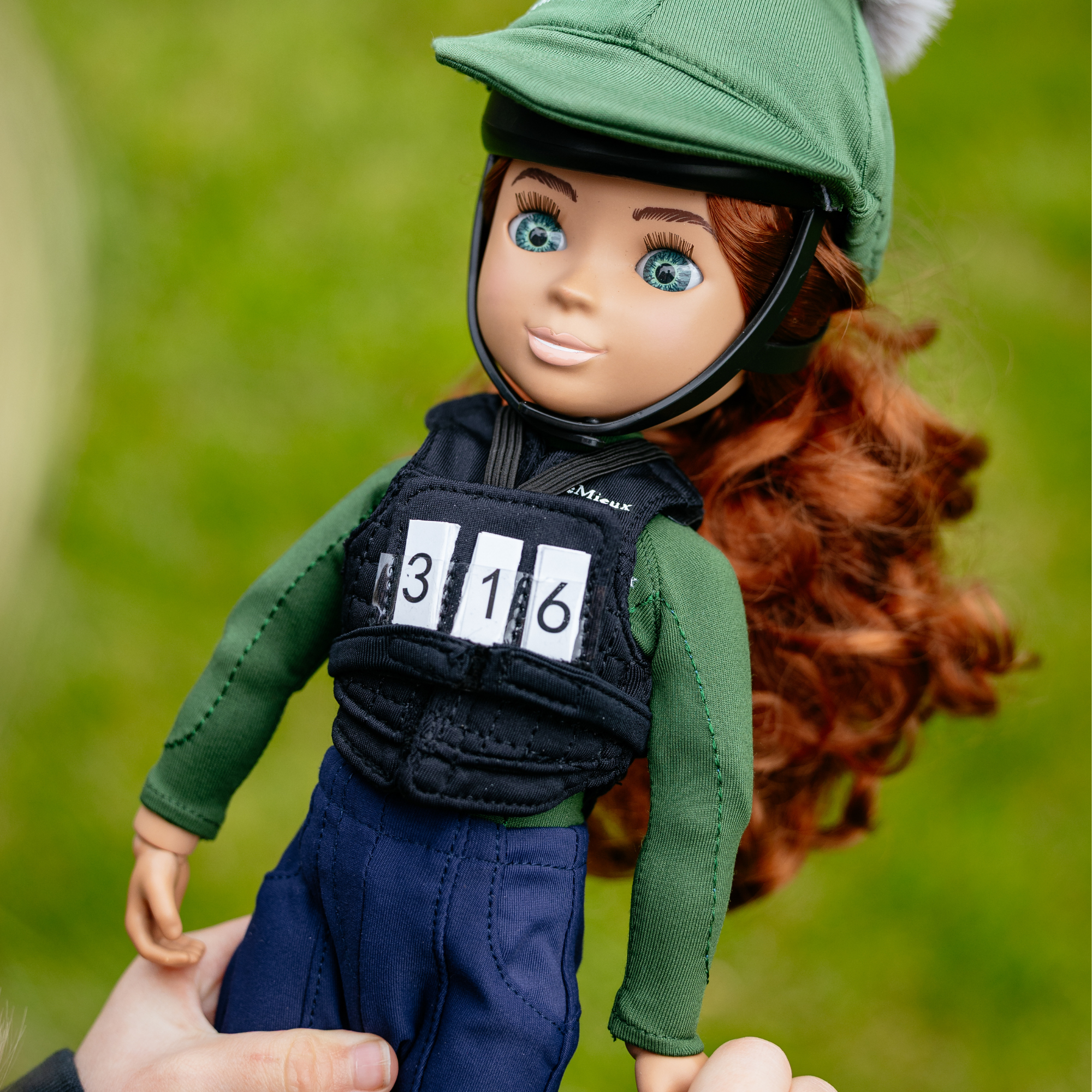 Doll in equestrian gear with a green helmet and dark vest, held against a blurred green background.