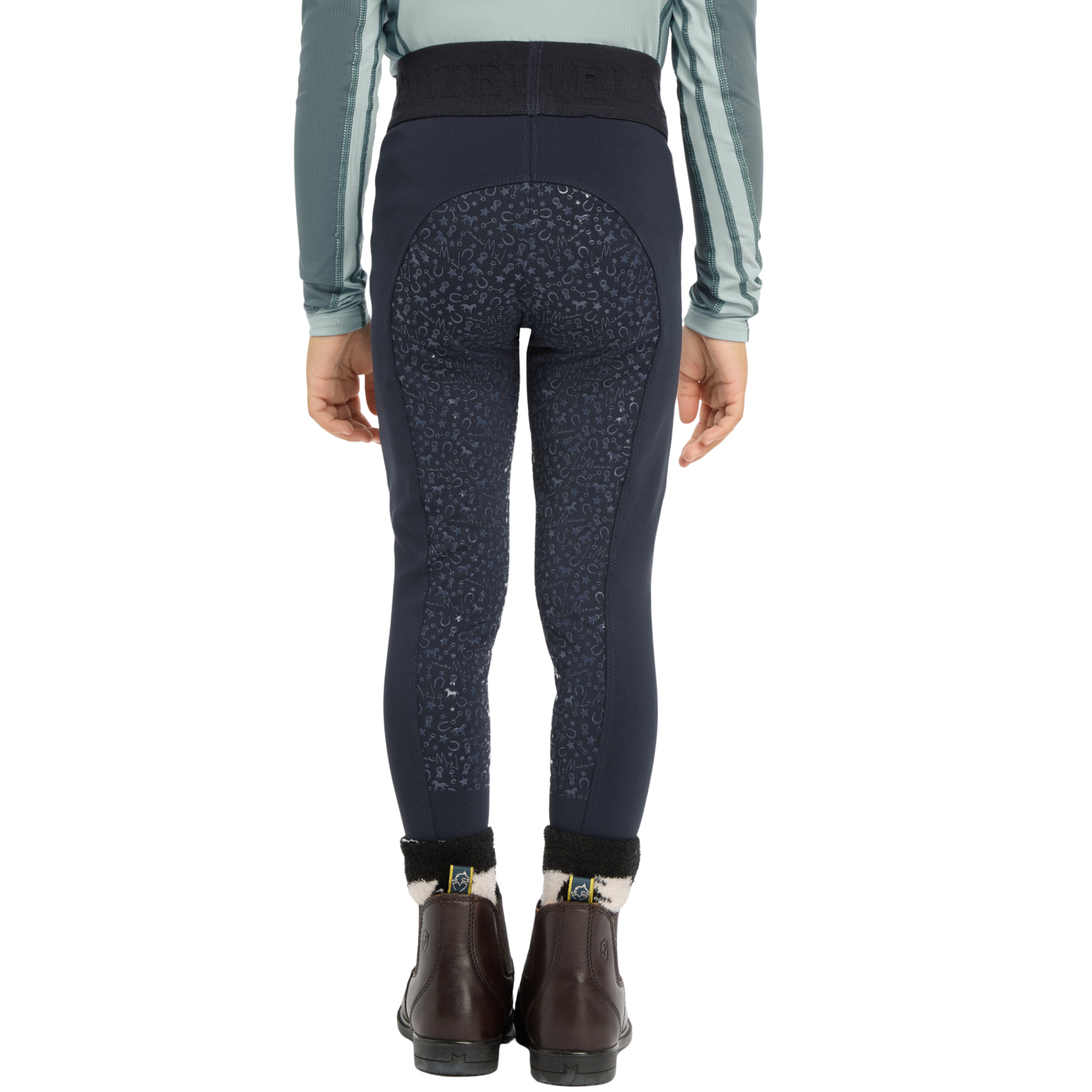 Rear view of a person wearing dark navy equestrian riding breeches with a subtle leopard pattern and a black waistband, paired with a sage green long-sleeve shirt and brown ankle boots, on a white background.