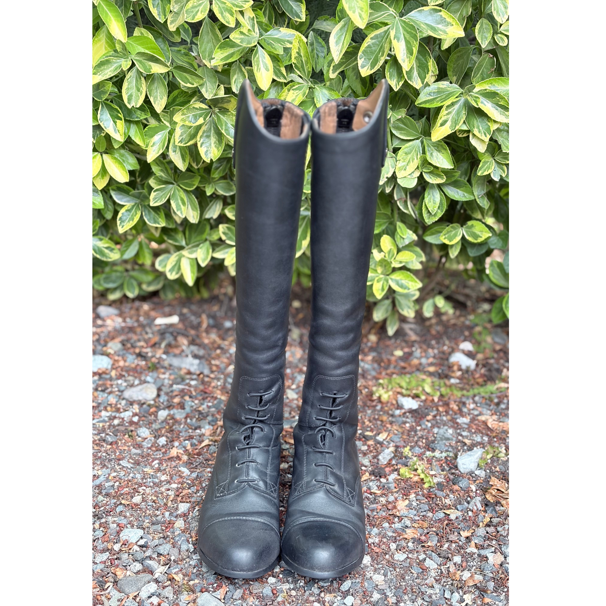 A pair of tall black leather riding boots with lace-up fronts, standing on a gravel surface in front of a green leafy hedge.