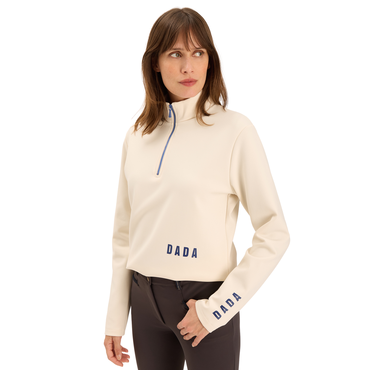 A woman modeling a cream-colored long-sleeve quarter-zip pullover sweatshirt with navy blue 'DADA' branding on the chest and sleeve, paired with dark brown pants against a solid white background.