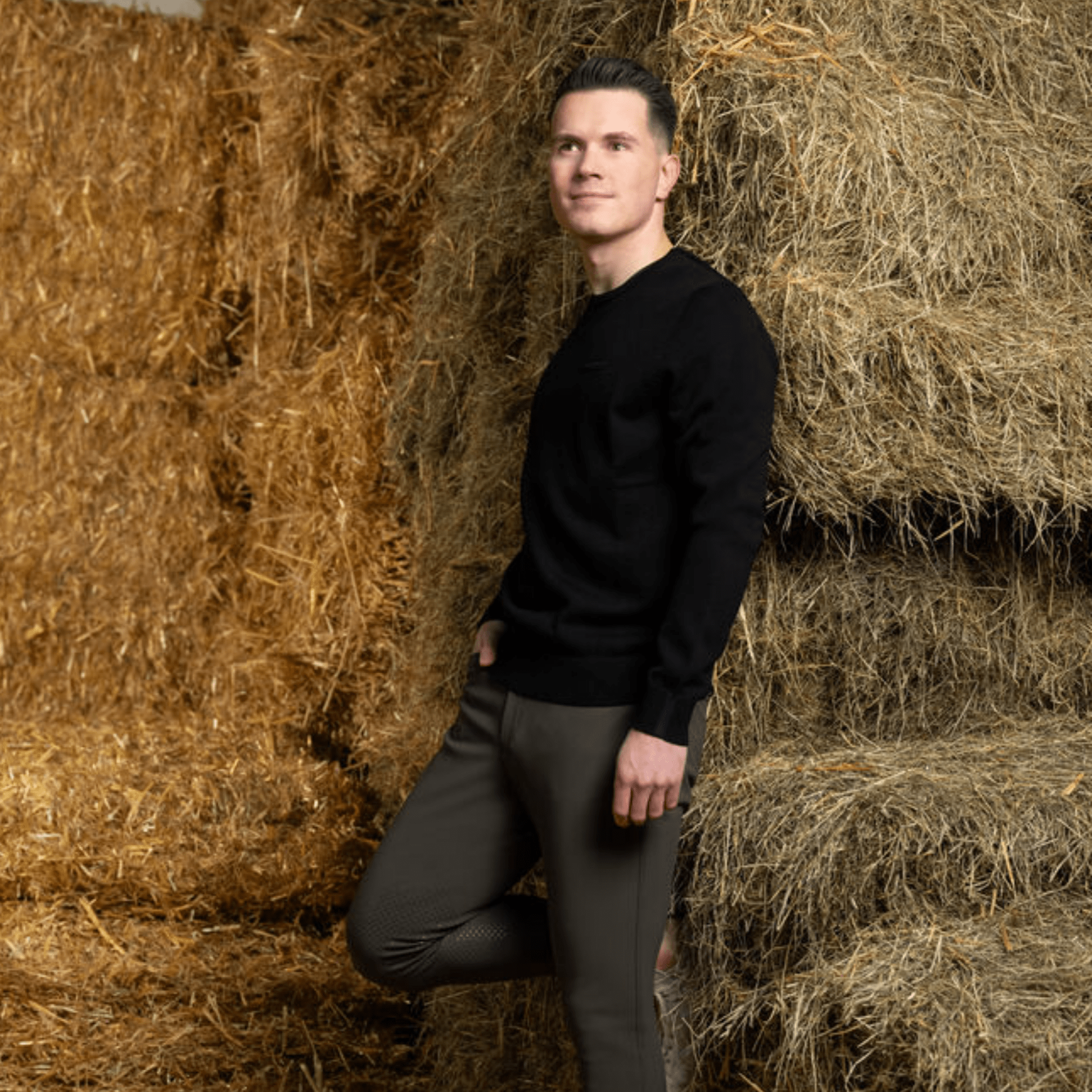 Man in a black sweater and green pants standing among hay bales.