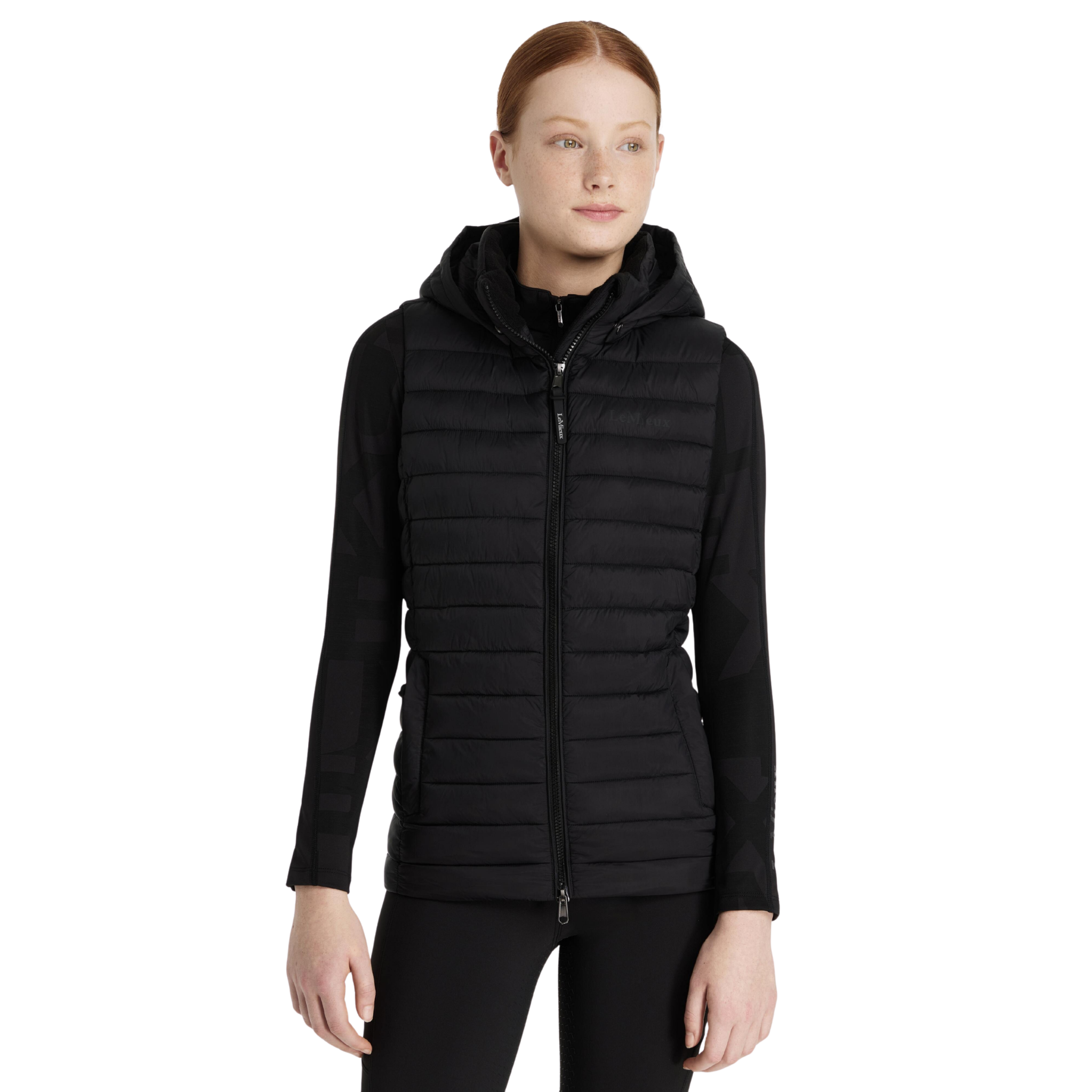 A woman modeling a black hooded zip-front puffer vest over a black top and black pants, photographed against a plain white studio background.