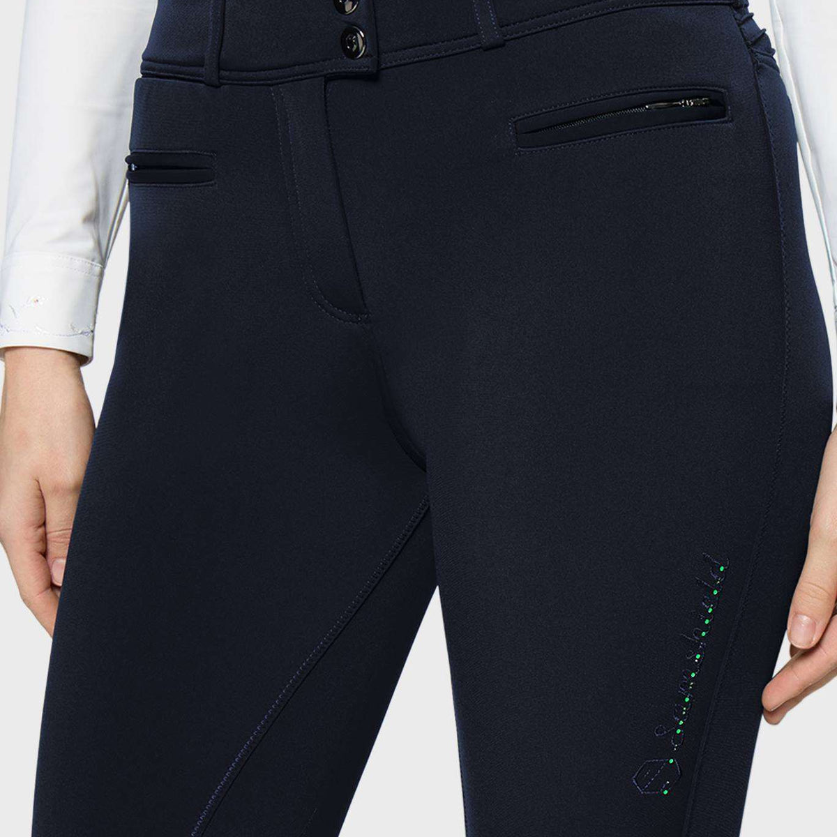 Close-up of a person wearing dark navy equestrian riding breeches, showing the front waistband with two buttons, side zip pocket, and leg stitching details.