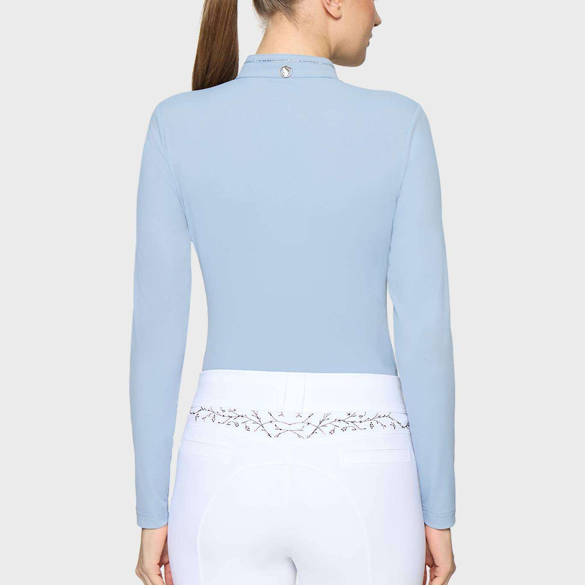 A woman shown from the back wearing a light blue long-sleeve mock-neck riding crop top and white riding breeches with a decorative silver floral waistband.
