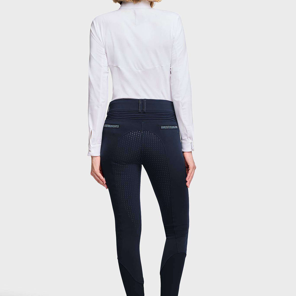 A woman shown from the back wearing a white long-sleeve shirt, dark navy textured riding breeches, and white sneakers against a light gray studio background.