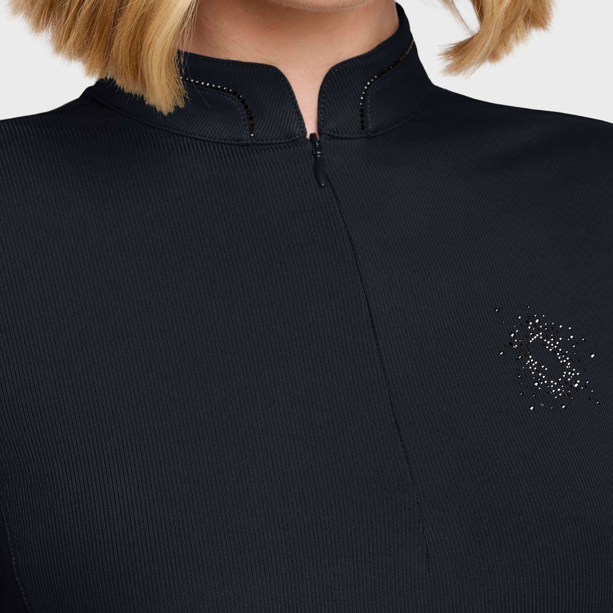 Close-up of a model wearing a black ribbed knit top with a mock neck, zipper, and a small rhinestone logo on the chest.
