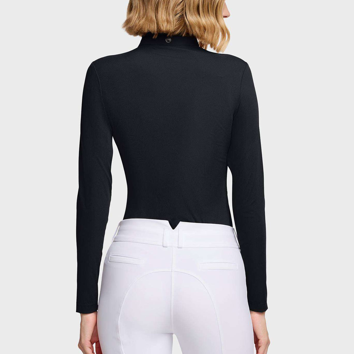 A woman shown from the back wearing a fitted black long-sleeve mock-neck top and white riding breeches against a plain white studio background.