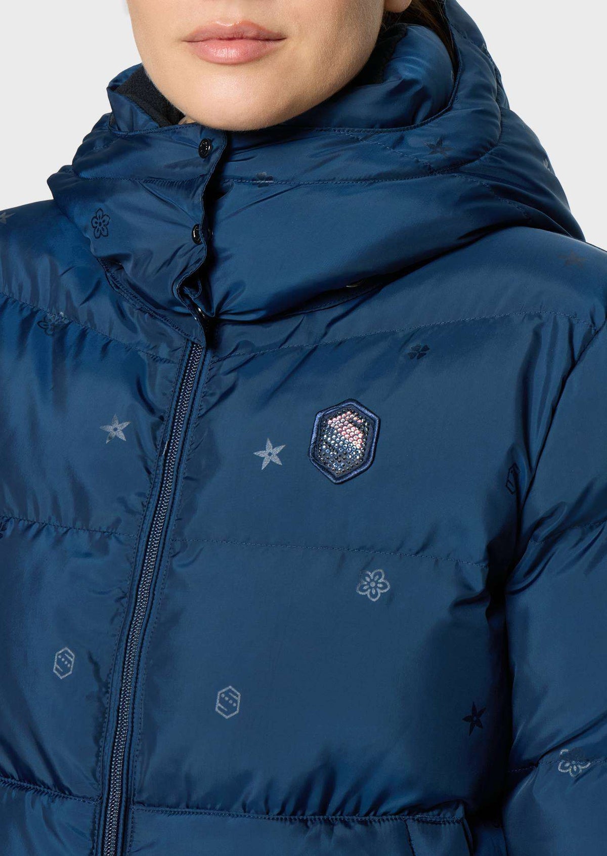 Close-up of a person wearing a navy blue hooded puffer jacket with a front zipper, quilted stitching, and small embroidered logo details on a light gray background.