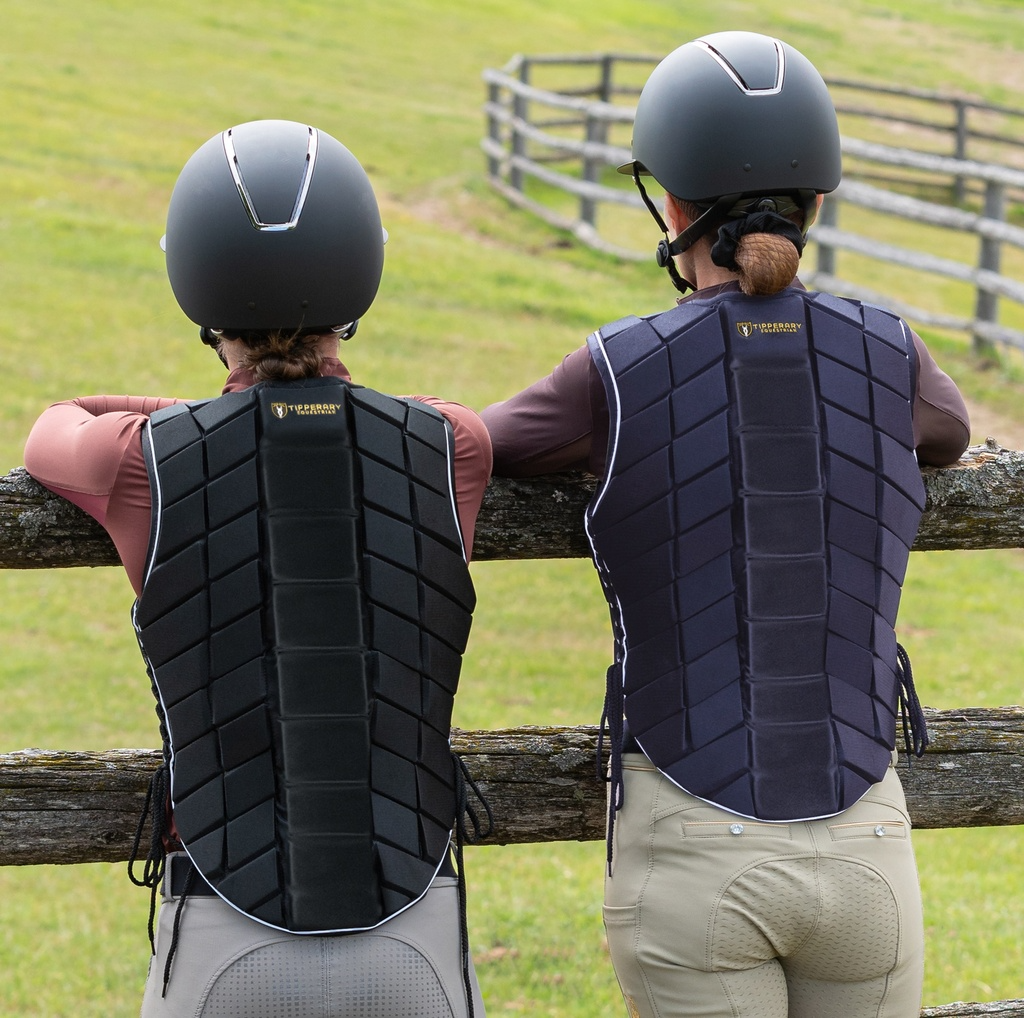 Two people wearing equestrian helmets and protective vests leaning against a wooden fence in an outdoor setting.