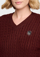 A close-up of a woman wearing a burgundy cable-knit V-neck sweater with a small hexagonal brooch on the chest.