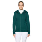 A woman modeling a dark green full-zip hoodie with a white turtleneck underneath and white leggings, set against a plain light gray studio background.