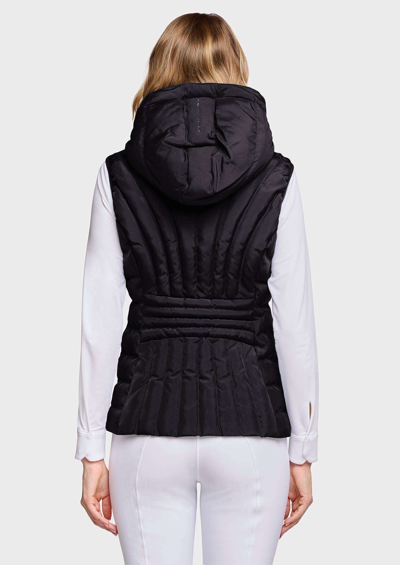 A woman viewed from the back wearing a black hooded puffer vest over a white long-sleeve top and white pants, against a light gray studio background.