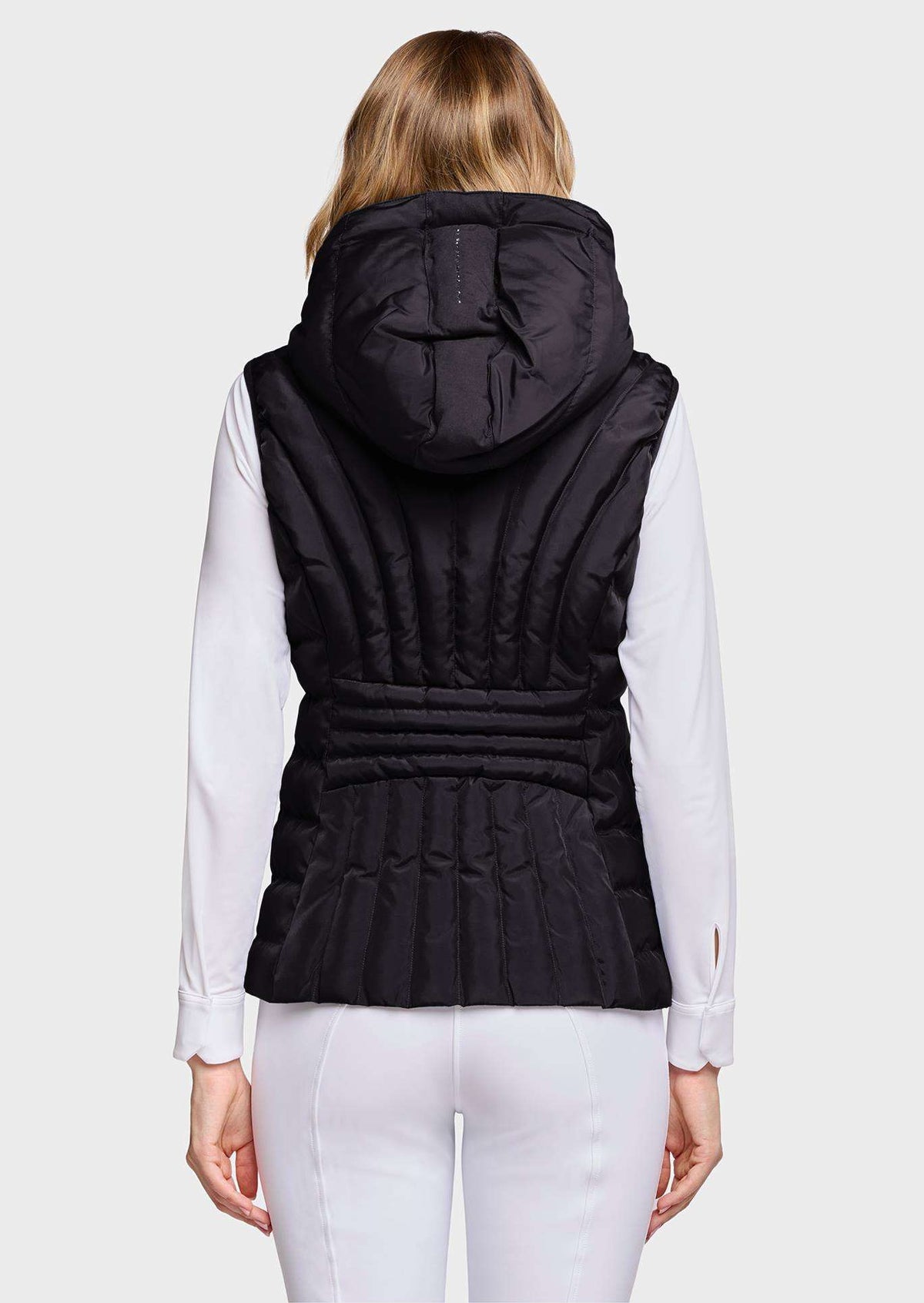 A woman viewed from the back wearing a black hooded puffer vest over a white long-sleeve top and white pants, against a light gray studio background.