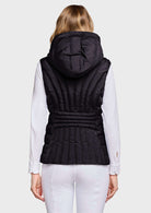 A woman viewed from the back wearing a black hooded puffer vest over a white long-sleeve top and white pants, against a light gray studio background.
