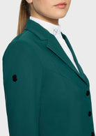 A close-up side view of a model wearing a dark teal green double-breasted blazer over a white mock neck top. The blazer features structured shoulders, a notched lapel, and black buttons, with a small black hexagonal logo patch on the upper sleeve.