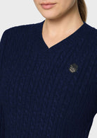 Close-up of a woman wearing a navy blue cable-knit V-neck sweater with a small circular embroidered logo on the chest, against a light gray background.