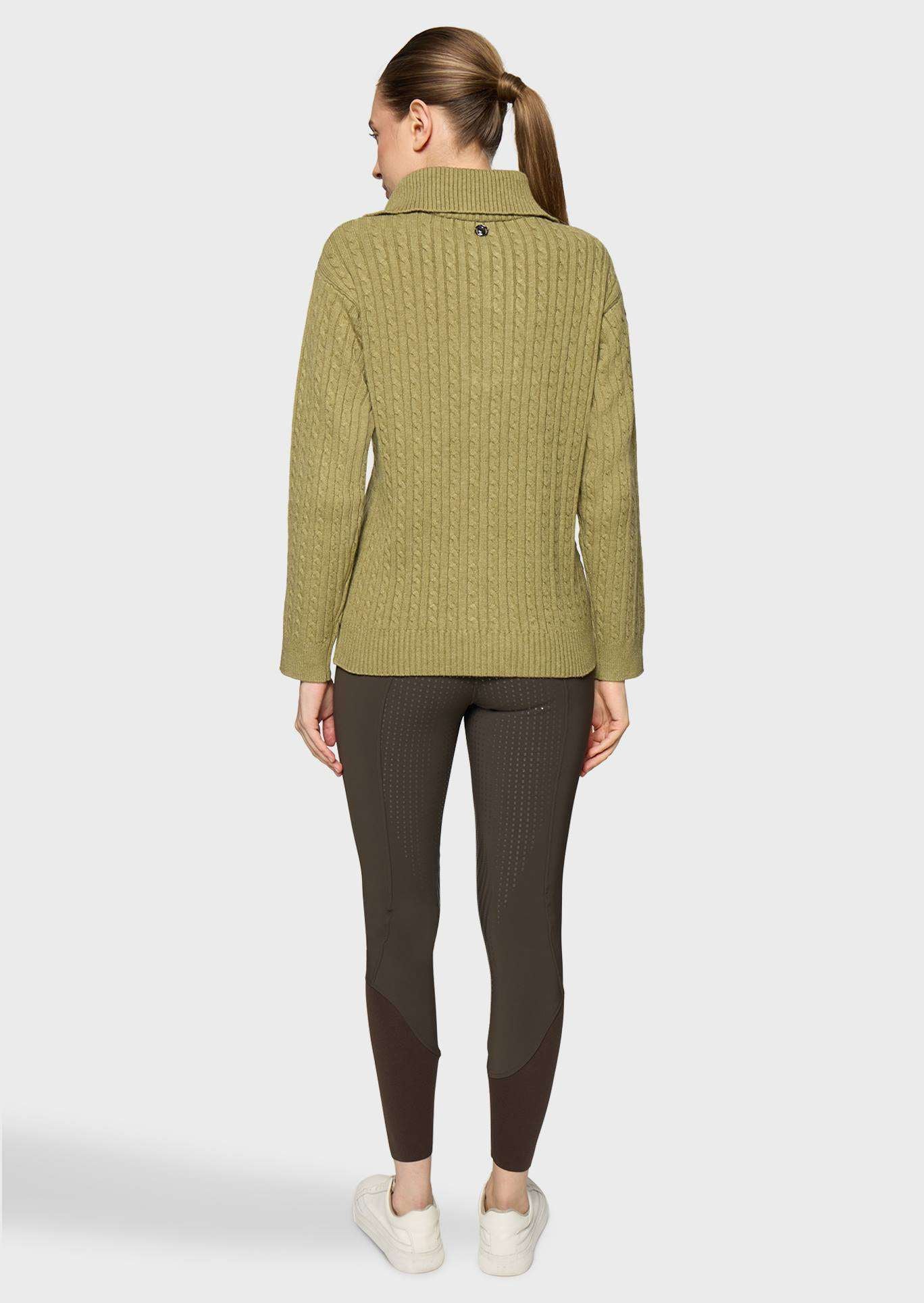A woman shown from the back wearing a textured olive green cable-knit zip-up sweater, dark brown ribbed leggings, and white sneakers against a light gray studio background.