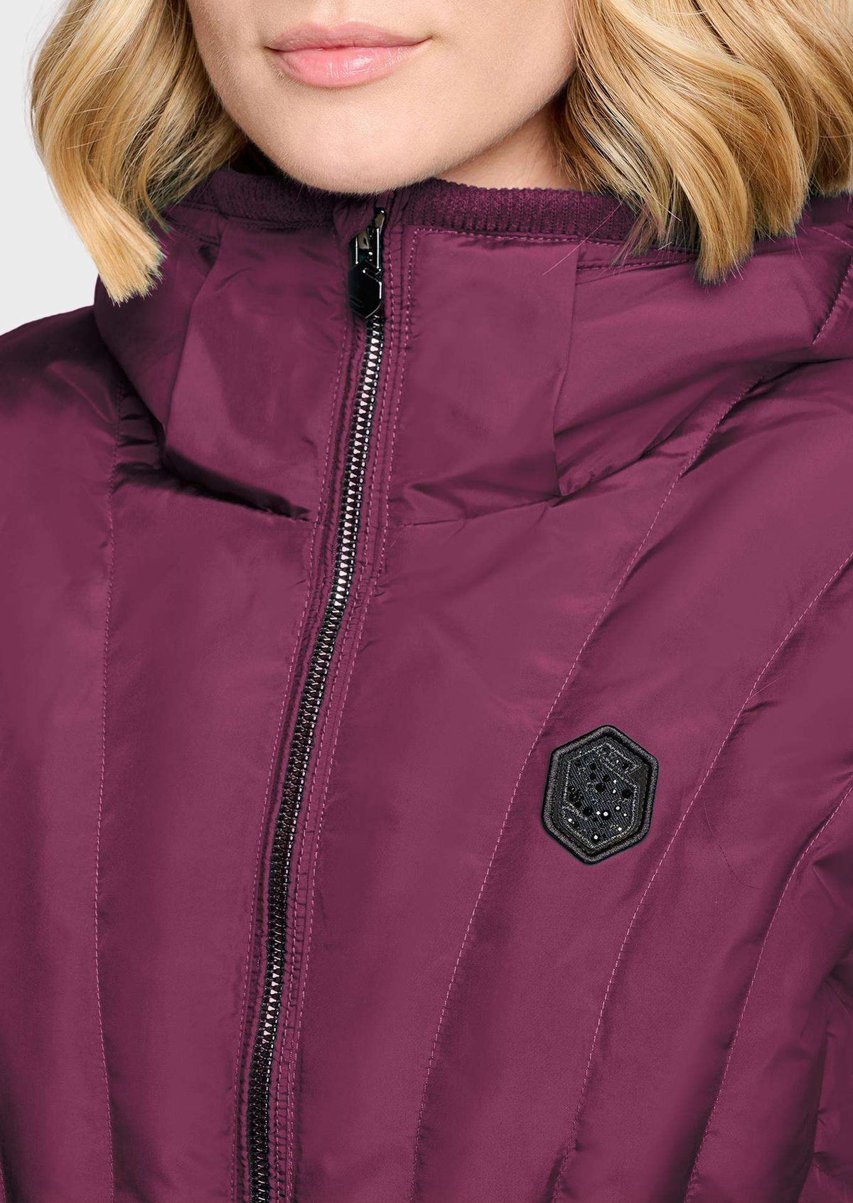 Close-up of a person wearing a quilted puffer jacket in deep plum purple. The jacket features a high ribbed collar, a front zipper, and a black hexagonal patch on the chest.