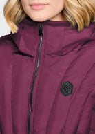 Close-up of a person wearing a quilted puffer jacket in deep plum purple. The jacket features a high ribbed collar, a front zipper, and a black hexagonal patch on the chest.