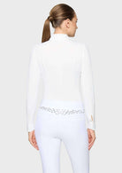 A woman shown from the back wearing a white long-sleeve mock-neck equestrian-style top and matching white fitted pants with a decorative silver embroidered waistband.