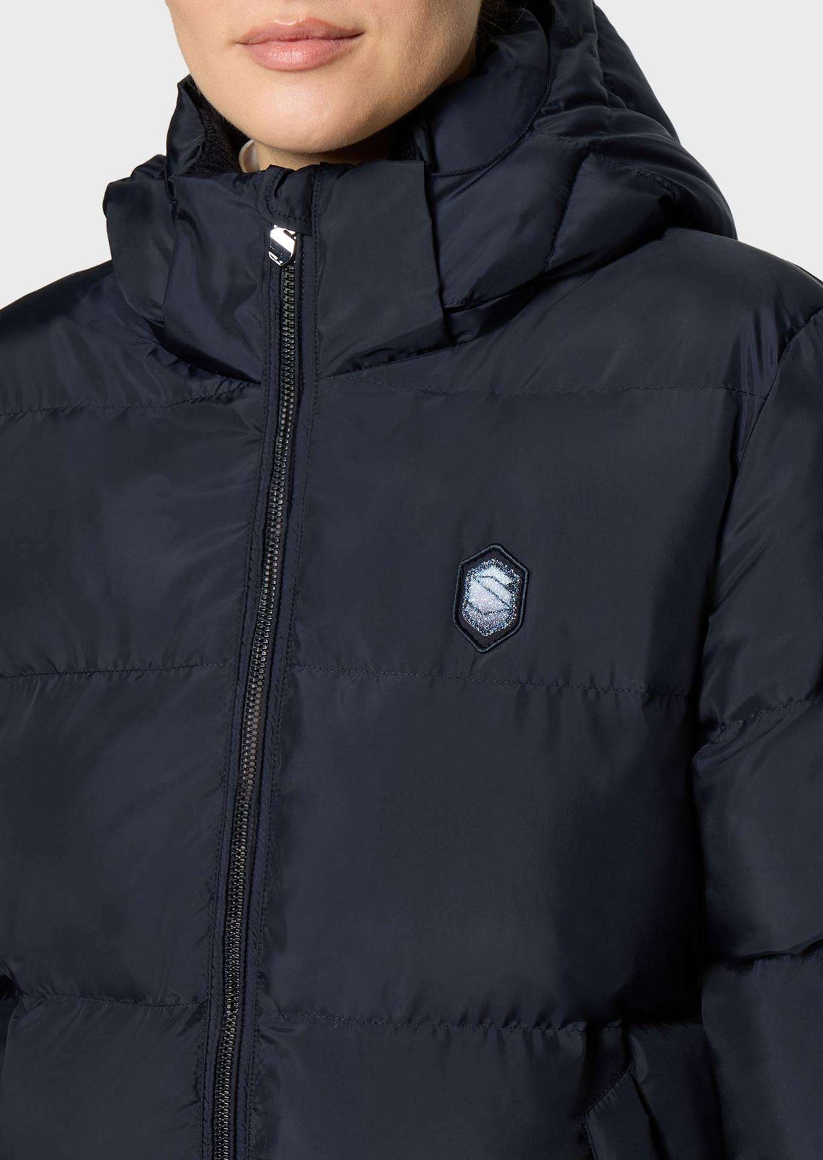Close-up of a person wearing a dark navy hooded puffer jacket with a front zipper and a small shield-shaped chest logo patch.