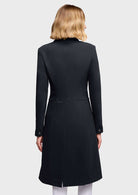 A woman shown from the back wearing a black longline coat with structured shoulders, long sleeves, and subtle embroidered detailing near the waist and hem, against a light gray studio background.