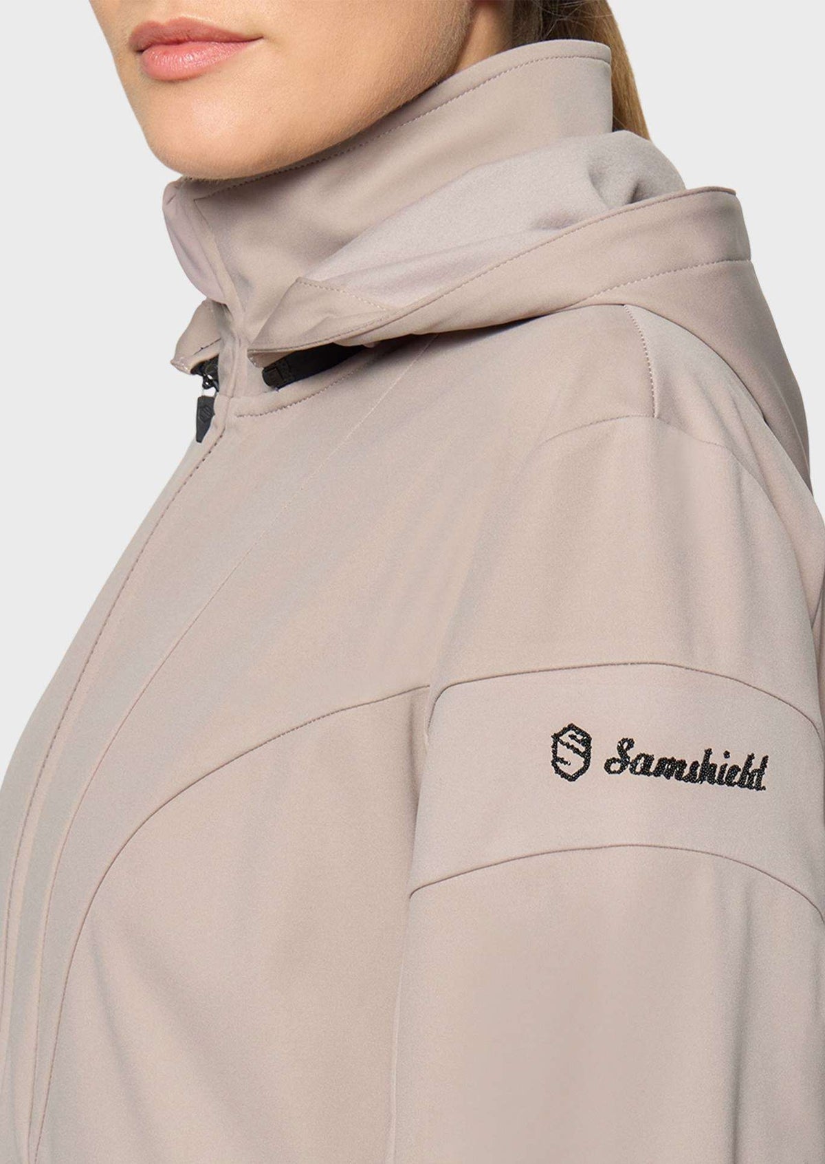 Close-up of a person wearing a light taupe hooded jacket with a black embroidered Samshield logo on the sleeve, against a plain light gray background.