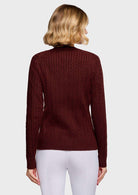 A woman viewed from the back wearing a burgundy cable-knit mock-neck sweater and white pants, against a light gray studio background.