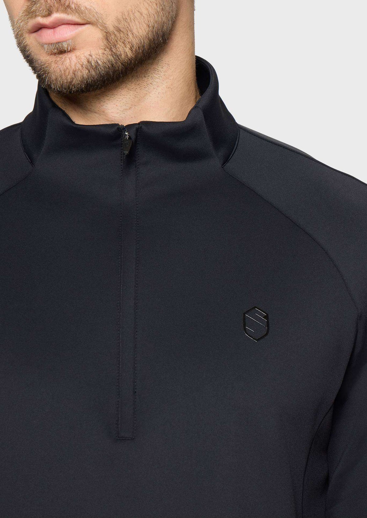 Close-up of a man wearing a black quarter-zip pullover with a small black geometric logo on the left chest, set against a light gray background.