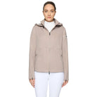 A woman modeling a light taupe hooded jacket with a full front zipper, zippered hand pockets, and zip cuffs, worn over a white turtleneck and white pants, against a light gray studio background.