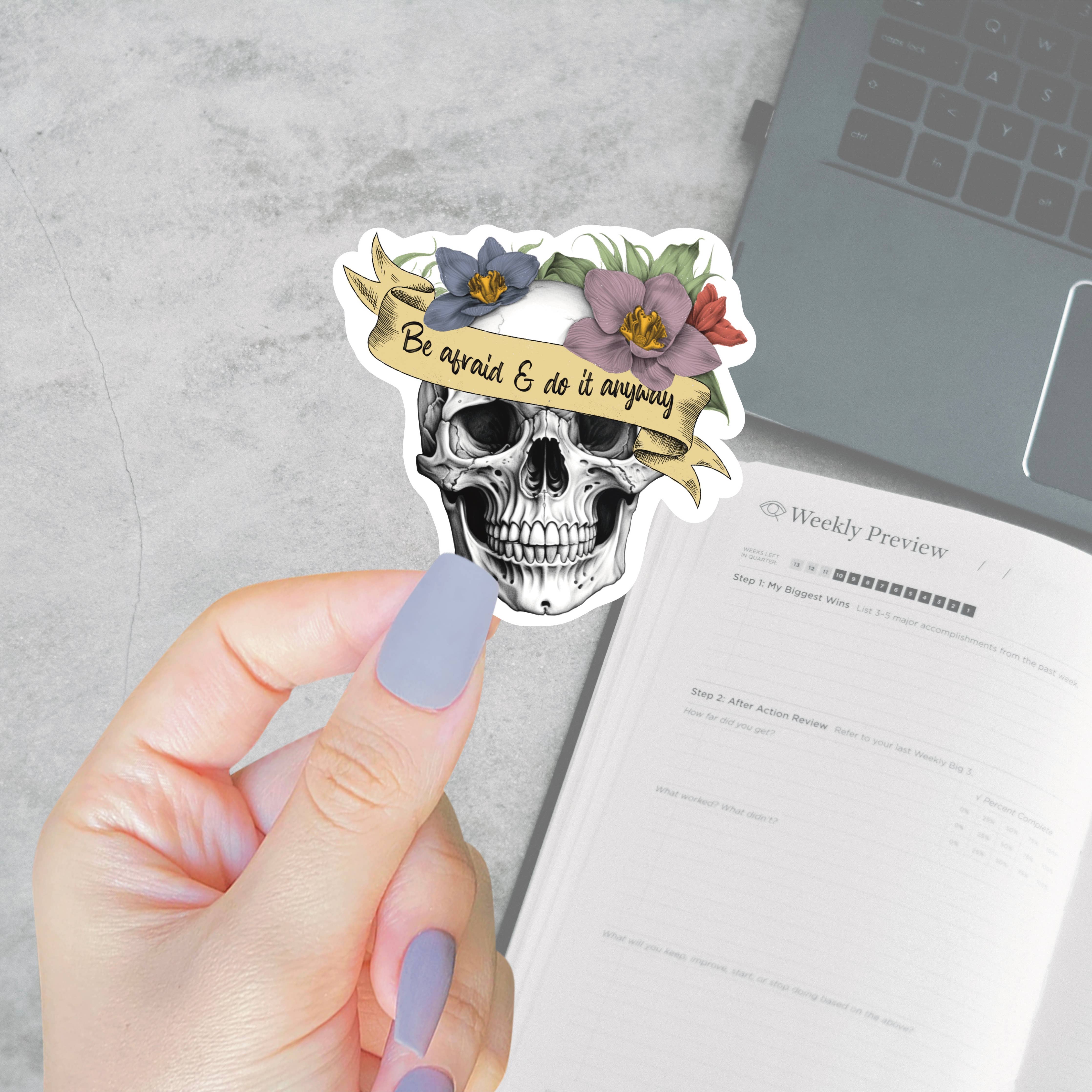 Skull sticker with floral crown held by a hand on a desk with a laptop and paper.