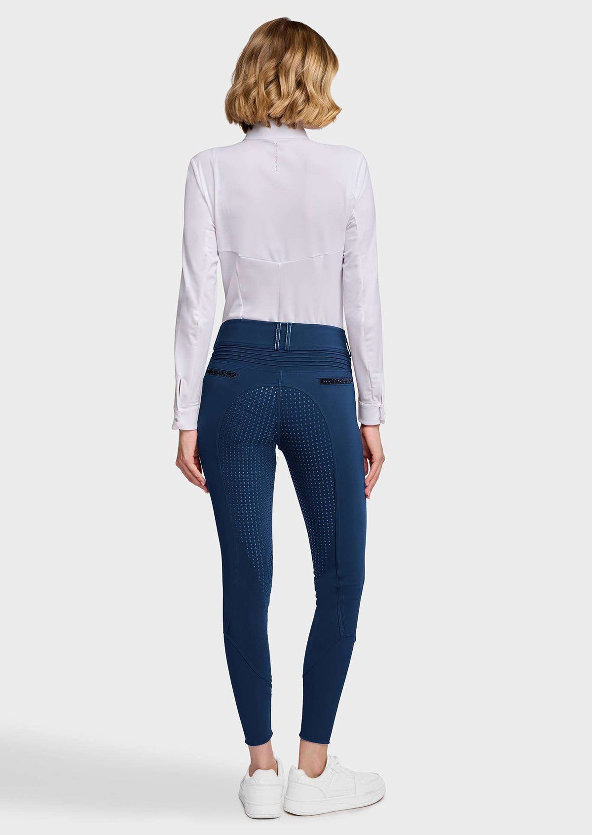 A woman shown from the back wearing a white long-sleeve shirt, navy blue patterned riding breeches, and white sneakers against a light gray studio background.