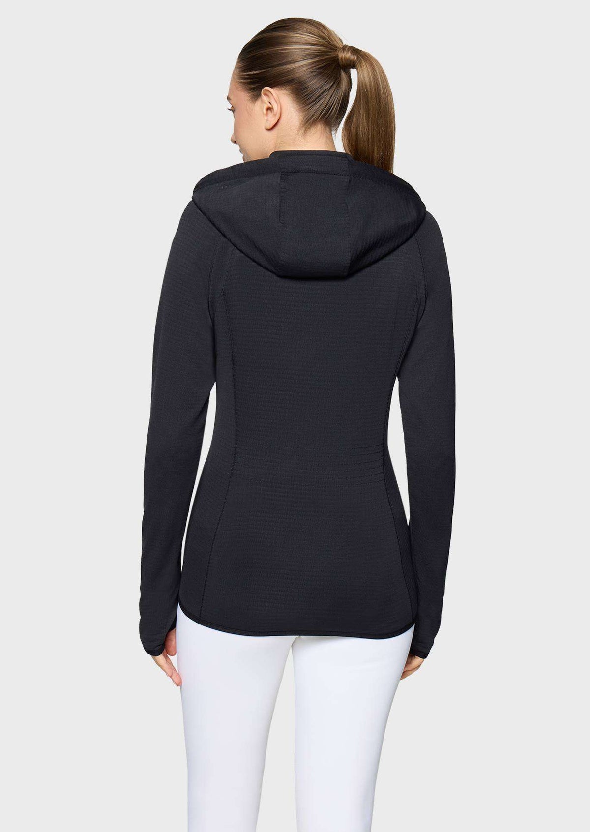 A woman viewed from the back wearing a black long-sleeve hooded athletic top and white leggings against a light gray studio background.