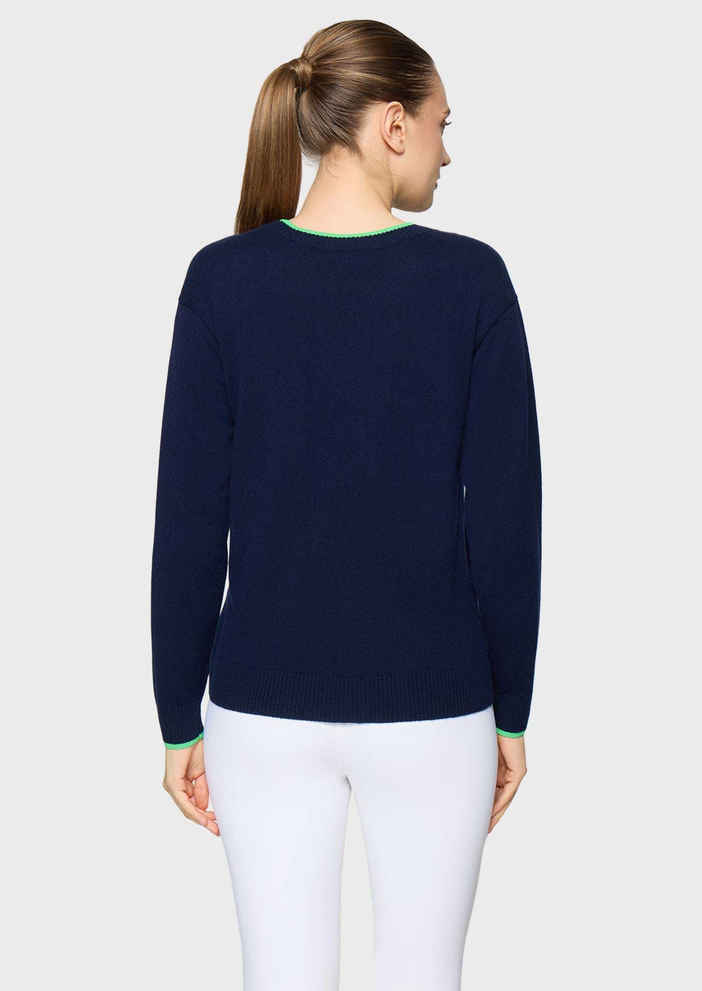 A woman viewed from the back wearing a navy blue knit sweater with light green trim at the crew neck and cuffs, paired with white pants, against a light gray studio background.