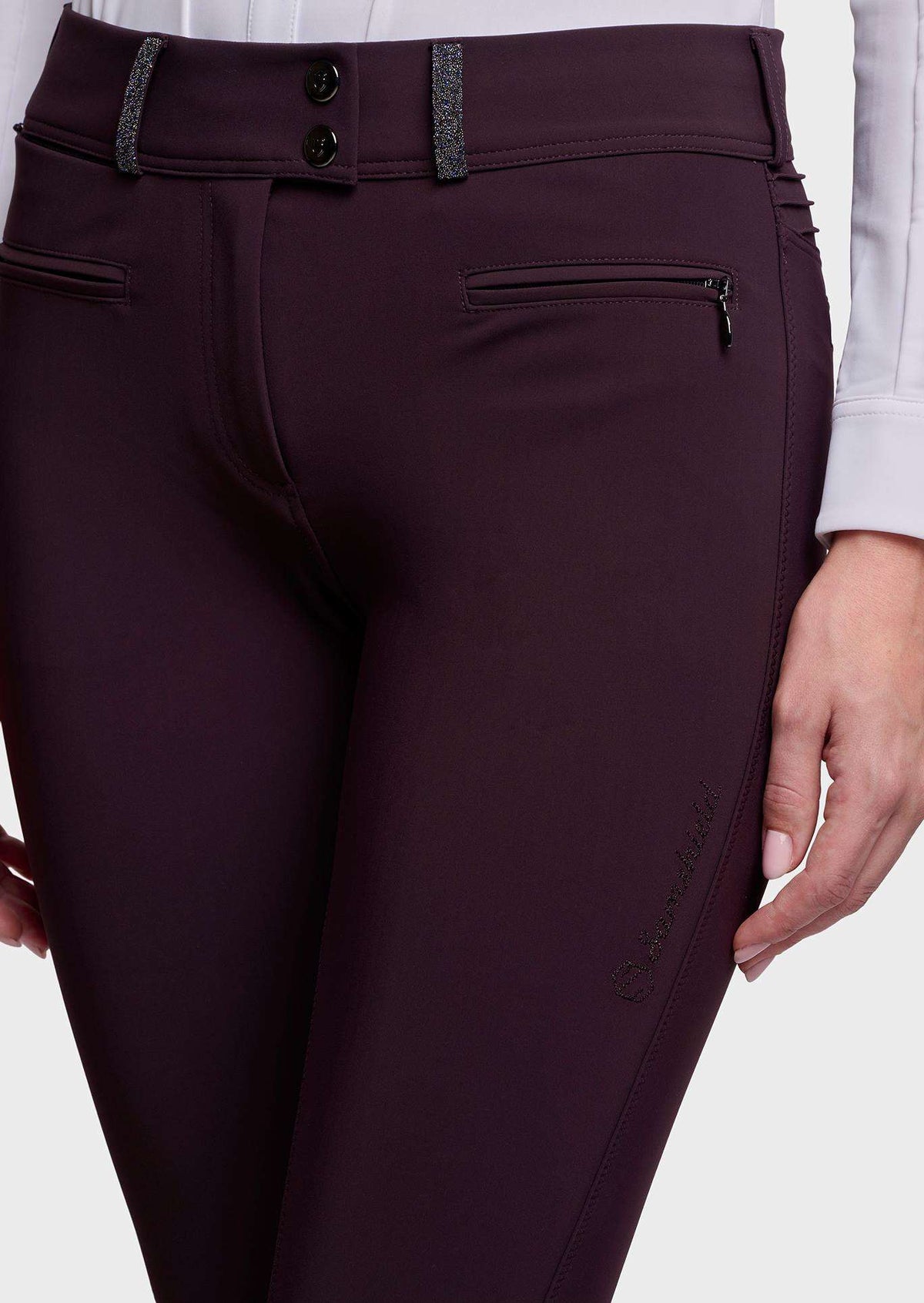 Close-up of a person wearing dark plum equestrian riding pants with a zippered pocket and decorative dark gray textured trim at the waistband, paired with a white long-sleeve shirt.