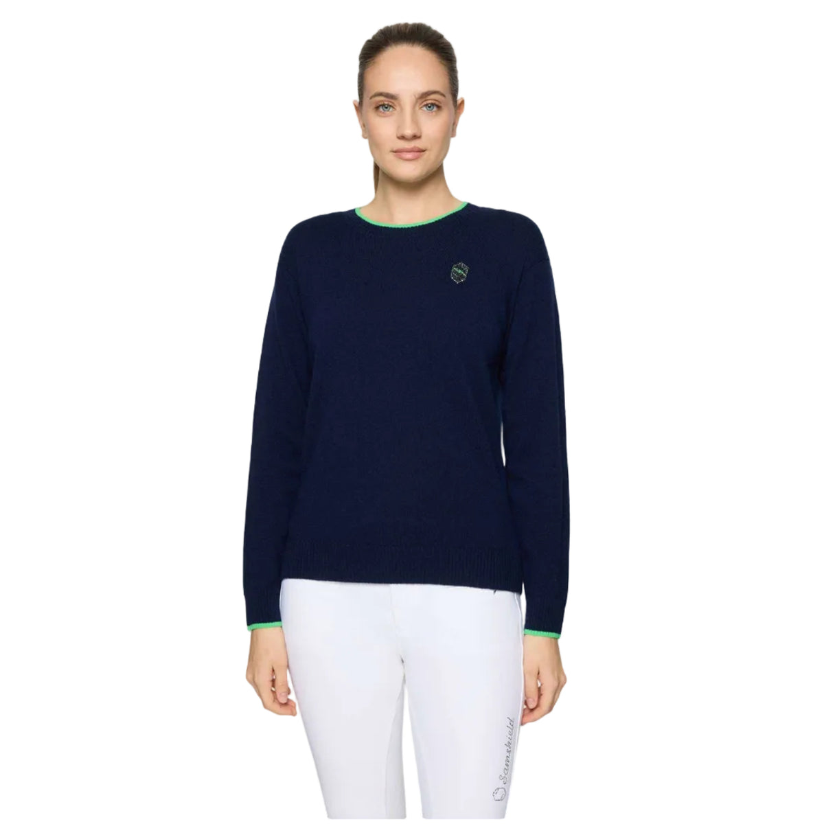 A woman modeling a navy blue crewneck sweater with bright green trim at the collar and cuffs, paired with white pants, against a light gray studio background.