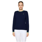 A woman modeling a navy blue crewneck sweater with bright green trim at the collar and cuffs, paired with white pants, against a light gray studio background.
